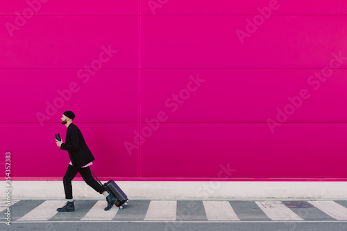 Young man with trolley walking along a pink wall, holding reusable cup