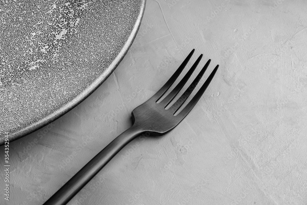 A black frosted fork lies next to a gray plate with the texture of concrete. A black frosted fork lies next to a gray plate with the texture of concrete.