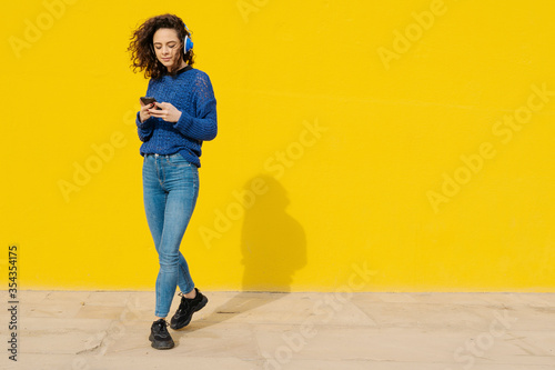 Portrait of young woman with headphones in front of yellow background looking at cell phone