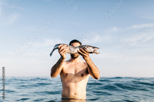 Young man standing in the sea holding caught fish in front of his face