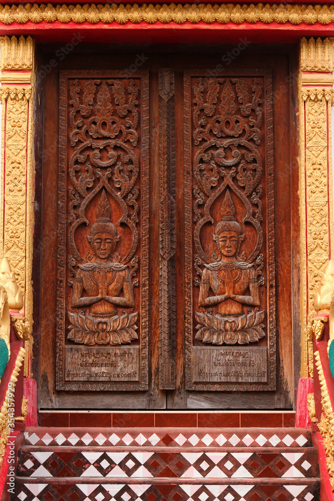 Entrance door with wood carvings showing praying buddha images in a temple site in Siamese Lao PDR, Southeast Asia