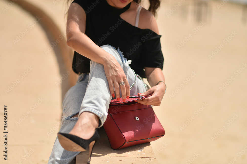 Opening a woman's bag. Sexy young woman on black t-shirt, fashion sunglasses and blue jeans opens up a red fashion bag.