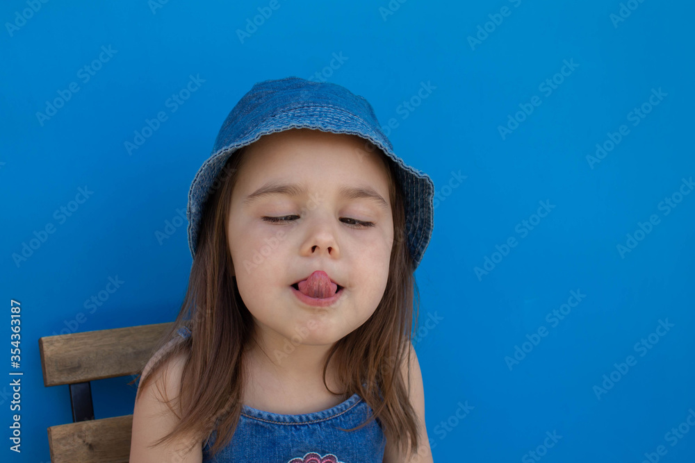 A girl in a Panama hat grimaces and shows her tongue against a blue isolated background A girl in a Panama hat grimaces and shows her tongue against a blue isolated background