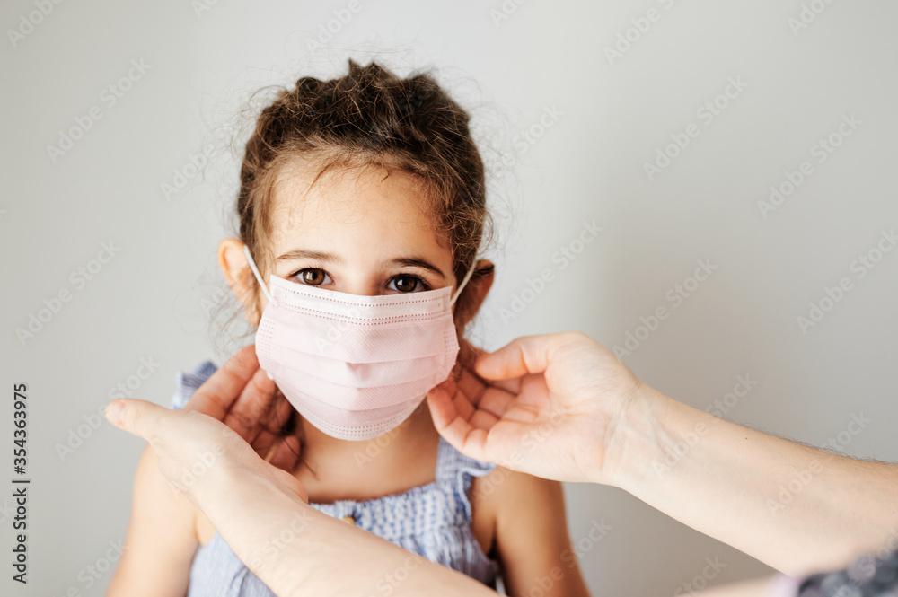 Girl with coronavirus mask prepared to go outside. Mother's hands putting the mask on her girl to prevent contagion Girl with coronavirus mask prepared to go outside. Mother's hands putting the mask on her girl to prevent contagion