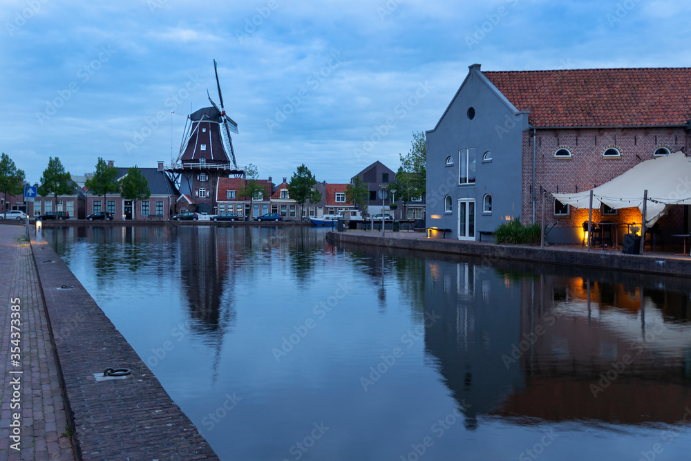 Obraz premium Holland - Meppel - a windmill in the middle of the town of Meppel, standing on the edge of a canal. The mill stands between the houses and is reflected in the water surface. The sky is with clouds