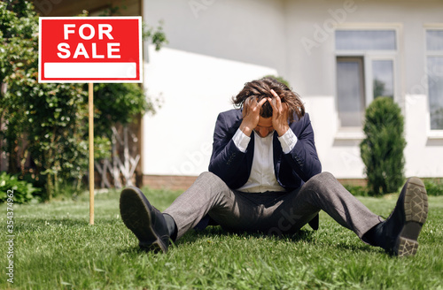 Obraz na plátne Depressed real estate agent sitting on grass in front of house for sale, outdoor