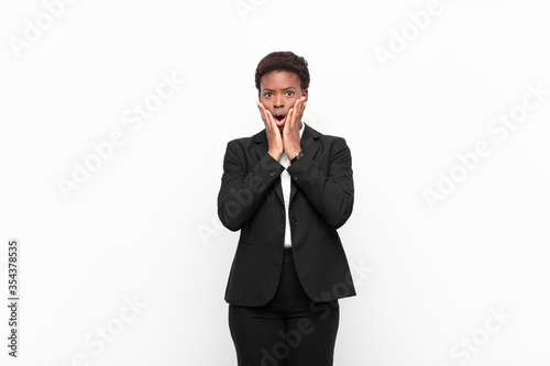 young pretty black womanfeeling shocked and scared, looking terrified with open mouth and hands on cheeks against white wall photo