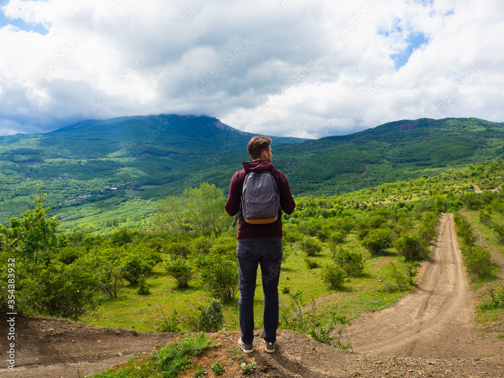 A young guy in a red hoodie and gray backpack travels in the mountains among green trees and clouds.