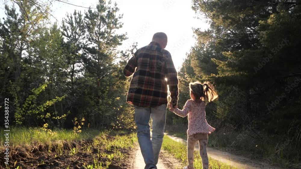 Rear view of old grandfather and little funny talking granddaughter walking along summer forest path in sunshine holding hands. Spend leisure time together outdoor for carefree lovely family happiness