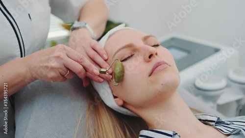 Close-up shot of facial massage with a jade roller on woman's face at beauty salon. Cosmetologist doing procedure for young woman.