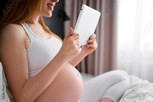 Technology and pregnancy. Young pregnant  woman using tablet computer sitting on a bed at home.