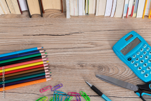 The new school and office supplies lie on a wooden desk top next to a row of books.