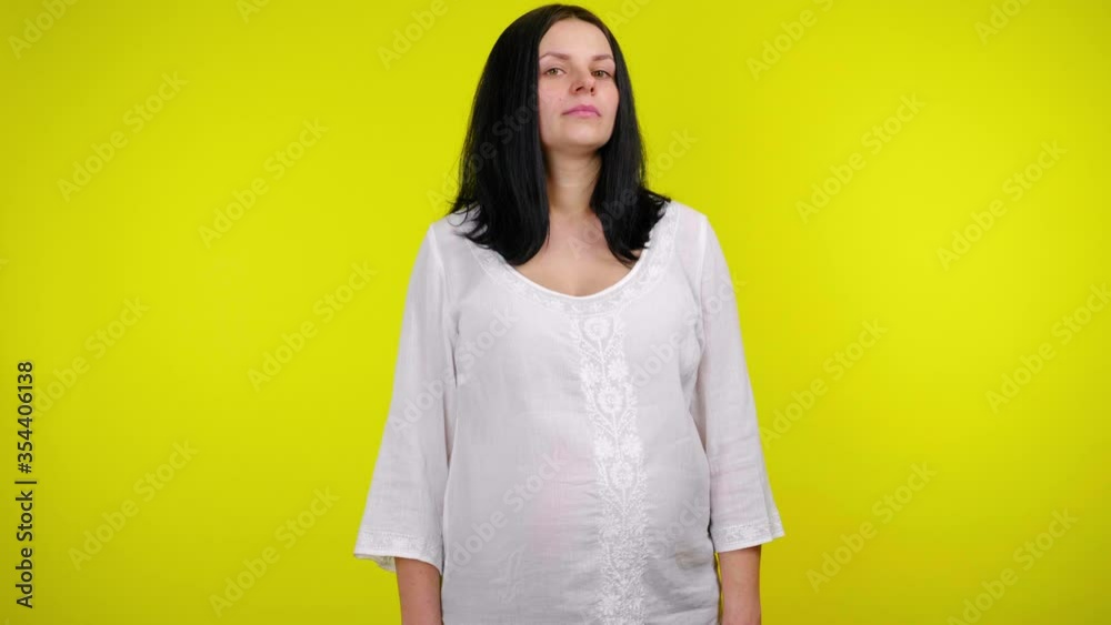 Young pregnant woman with black hair in a white blouse looks around on a yellow background with copy space. Place for text or product. 4K slow motion footage Young pregnant woman with black hair in a white blouse looks around on a yellow background with copy space. Place for text or product. 4K slow motion footage
