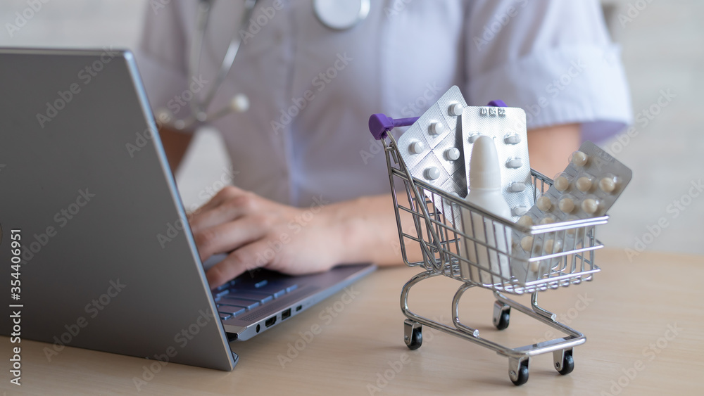 Unrecognizable female doctor prints a report on a laptop. Online pharmacy concept. The faceless pharmacist sells drugs on the Internet. Mini shopping trolley beatery tablets.