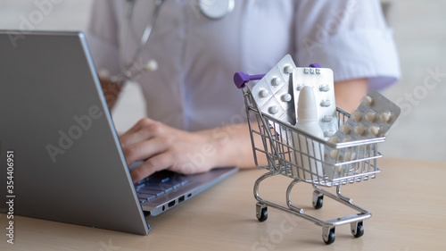 Unrecognizable female doctor prints a report on a laptop. Online pharmacy concept. The faceless pharmacist sells drugs on the Internet. Mini shopping trolley beatery tablets.