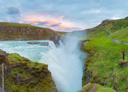 Fototapeta Naklejka Na Ścianę i Meble -  Amazing summer sunrise on Gullfoss Waterfall.
A man enjoys a view of a waterfall standing on a cliff. Colorful morning scene of Iceland. Midnight sun of Iceland. Visit Iceland. Beauty world.