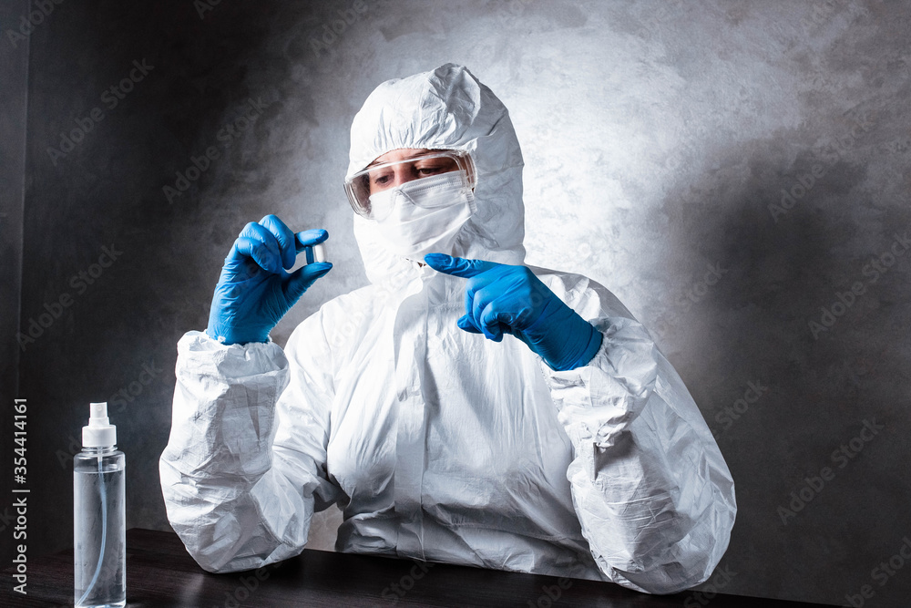 Nurse in a medical face mask and a viral protective suit. The doctor is sitting at the table in a medical face mask and a viral protective suit. The doctor holds a pill in his hands.