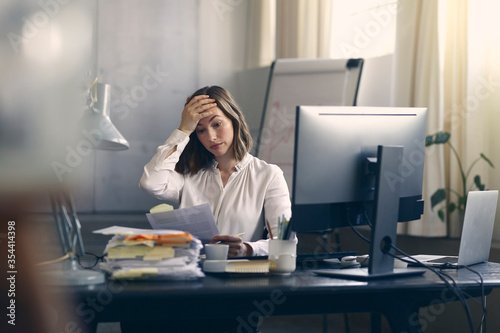 Very stressed business woman sitting in front of her computer looking at a large pile of paperwork, while holding a hand at her forehead  photo