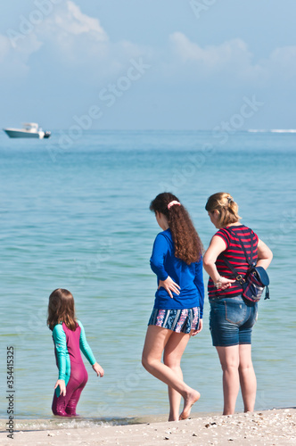 Back view, medium distance of a mother and daughter watching another daughter playing on the shoreline of tropical, sandy beach on gulf of Mexico on a sunny day the 