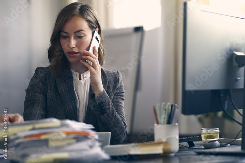 Beautiful brunette business woman and  female editor talking on the phone while looking down at some paper work at her office desk. Sitting in a modern office space. Concentrated about work. 