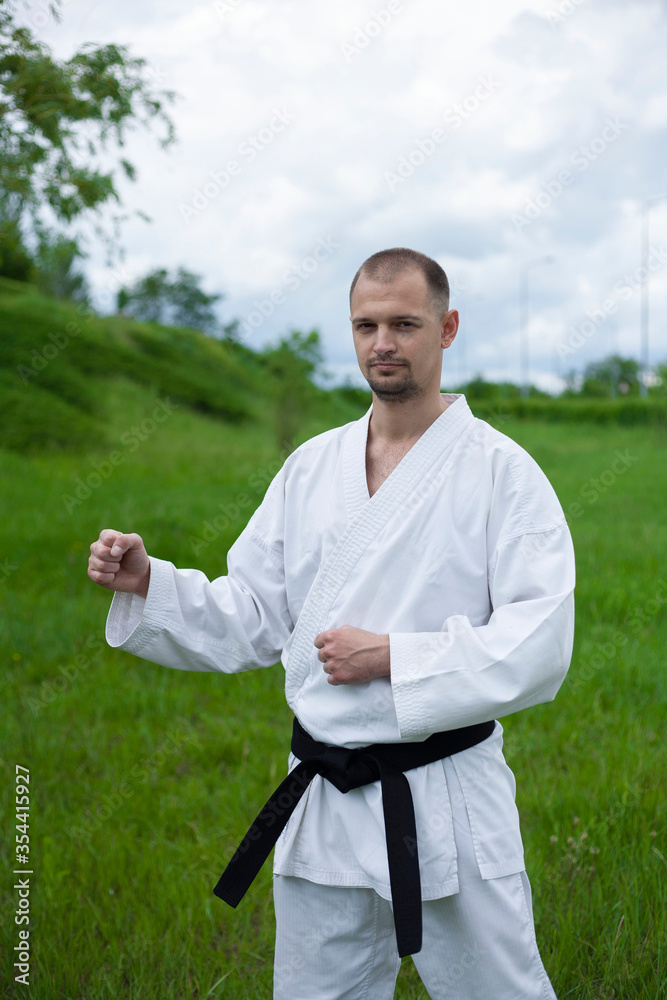 A karate trainer stands on the lawn and holds his hands in a protective pose. White kimono. Black belt in karate. Training in the fresh air.