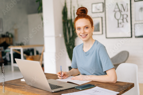 Successful redhead young woman is writing in documents, working on laptop while sitting at the desk in light cozy living room at home office, looking at camera. Concept of remote working.