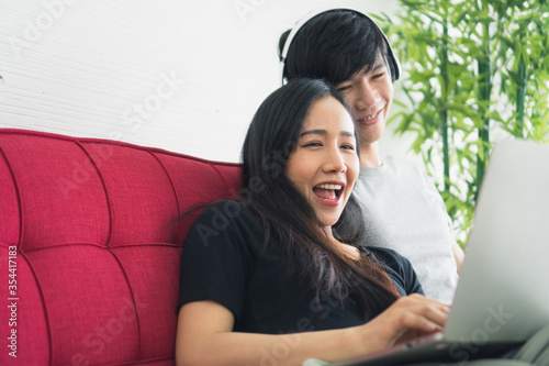 Young Asian couple love man and woman working at home by use computer laptop notebook sitting on red sofa in living room smiling and felling happy. corona virus, COVID-19 Pandemic new normal Concept