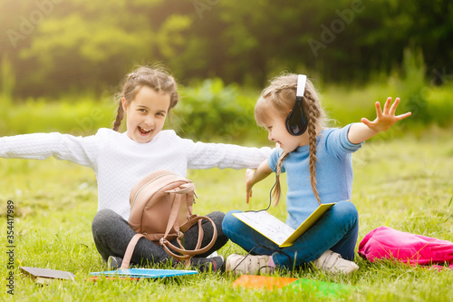 two cute multicultural schoolgirls sitting on lawn under tree and reading book together