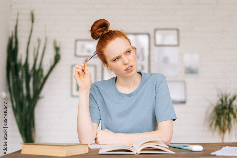Thoughtful redhead young woman student is making an outline from textbook in workbook at desk at home office. Thoughtful redhead young woman student is making an outline from textbook in workbook at desk at home office.