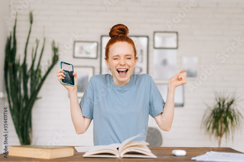 Happy redhead young woman is feeling euphoric reading good amazing news on cell phone while sitting at desk with paper books at home office. Concept of remote work or education from home.
