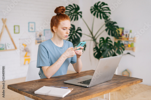 Serious pretty redhead young woman uses the phone while sitting at the desk in light cozy room at home office. Concept of remote working on distance workplace.