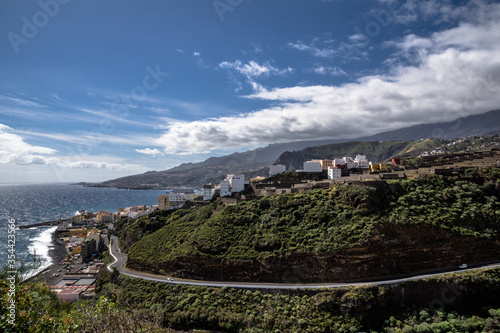 View of Santa Cruz and a coastal road  of La Palma. Steep and wooded mountainsides  on which houses are partly built  flat down to the sea . The blue sky is partly covers by white clouds.