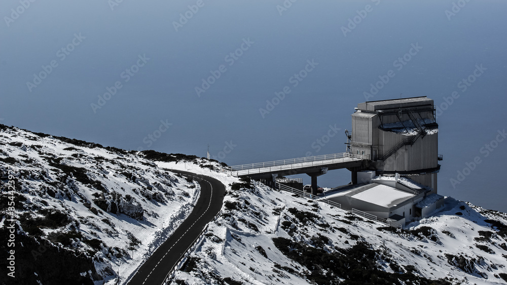 Obraz premium A abstrakt looking building of the observatory on the Canary Island La Palma. A road leads to the building. The ground is covered with snow and in the background the Atlantic Ocean is visible.