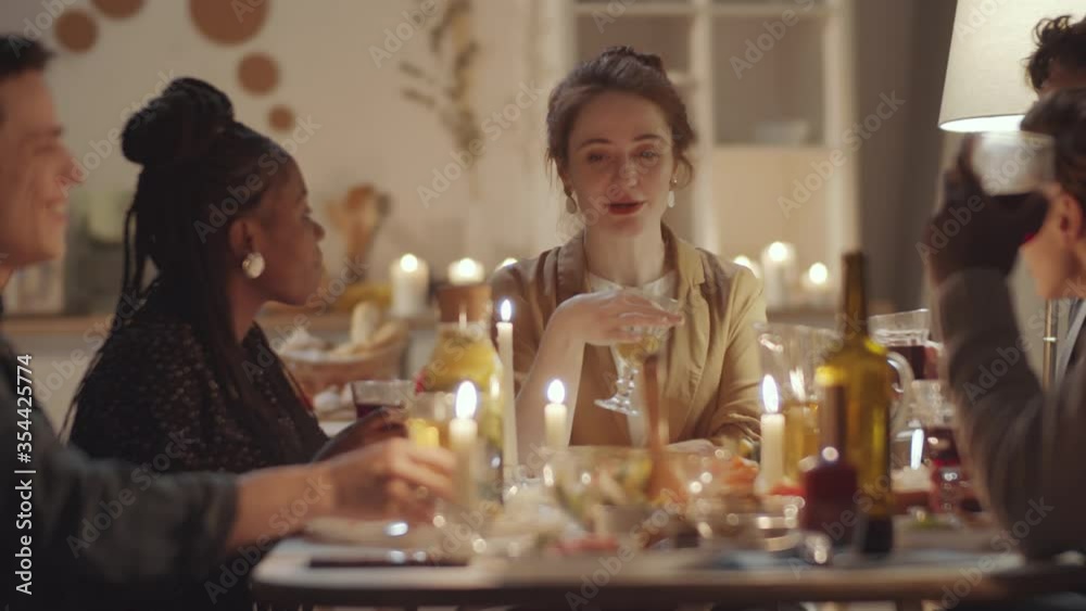 Group of multiethnic friends sitting together at dinner table with candles and food on it, clinking glasses together and drinking wine after cheerful woman giving a toast at home party