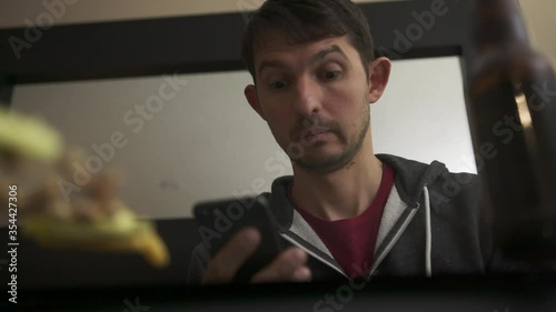 High quality slow motion close-up 4k video of a young blurred man chatting with friend or family on phone watching tv and having snacks with bottle of beer. Happy time. Handsome person with phone. 