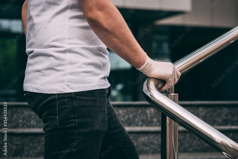 Male hand in a white latex medical glove on the railing..white T-shirt and black jeans..man wearing protective latex gloves touching railing outsid