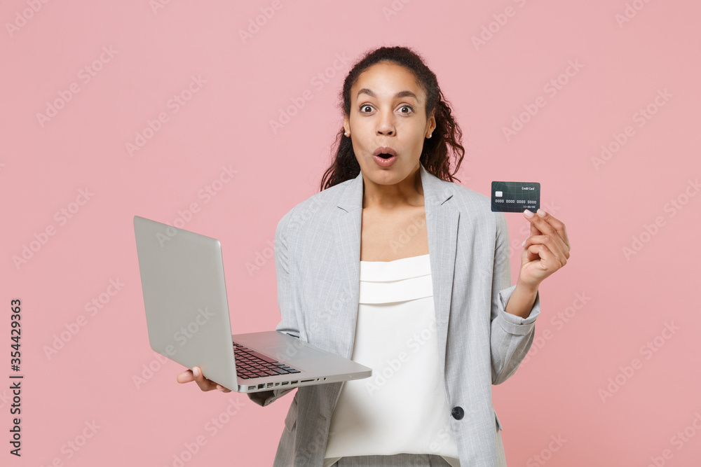 Shocked african american business woman in grey suit white shirt isolated on pink background. Achievement career wealth business concept. Mock up copy space. Hold laptop pc computer credit bank card. Shocked african american business woman in grey suit white shirt isolated on pink background. Achievement career wealth business concept. Mock up copy space. Hold laptop pc computer credit bank card.