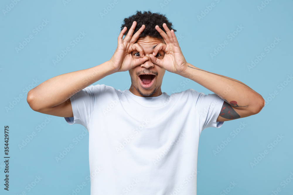 Excited young african american guy in casual white t-shirt posing isolated on blue background. People lifestyle concept. Mock up copy space. Holding hands near eyes, imitating glasses or binoculars. Excited young african american guy in casual white t-shirt posing isolated on blue background. People lifestyle concept. Mock up copy space. Holding hands near eyes, imitating glasses or binoculars.
