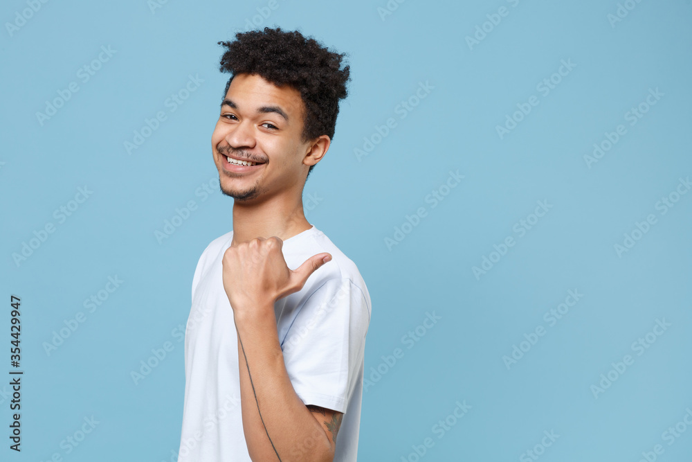 Side view of smiling young african american guy in casual white t-shirt posing isolated on pastel blue background studio portrait. People lifestyle concept. Mock up copy space. Pointing thumb aside. Side view of smiling young african american guy in casual white t-shirt posing isolated on pastel blue background studio portrait. People lifestyle concept. Mock up copy space. Pointing thumb aside.