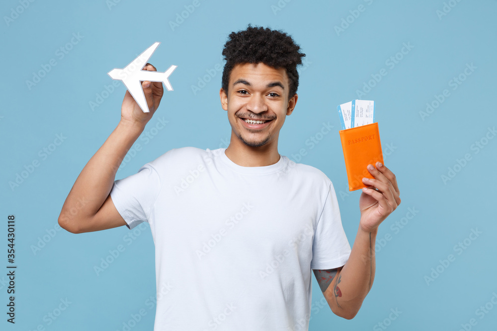 Smiling young african american guy in casual white t-shirt traveling abroad isolated on blue background. Air flight journey concept. Mock up copy space. Hold passport tickets boarding pass air plane. Smiling young african american guy in casual white t-shirt traveling abroad isolated on blue background. Air flight journey concept. Mock up copy space. Hold passport tickets boarding pass air plane.
