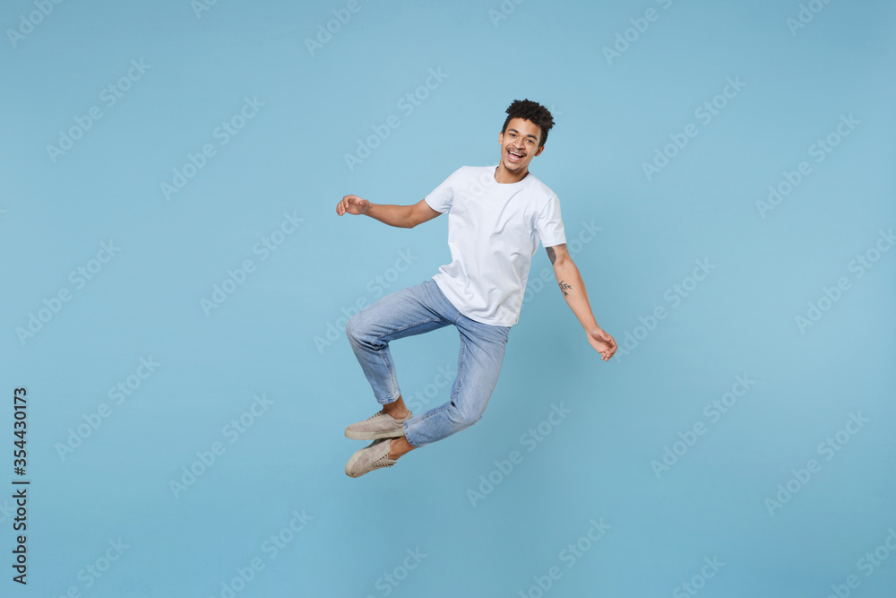 Cheerful young african american guy in casual white t-shirt posing isolated on pastel blue wall background studio portrait. People emotions lifestyle concept. Mock up copy space. Jumping, having fun.