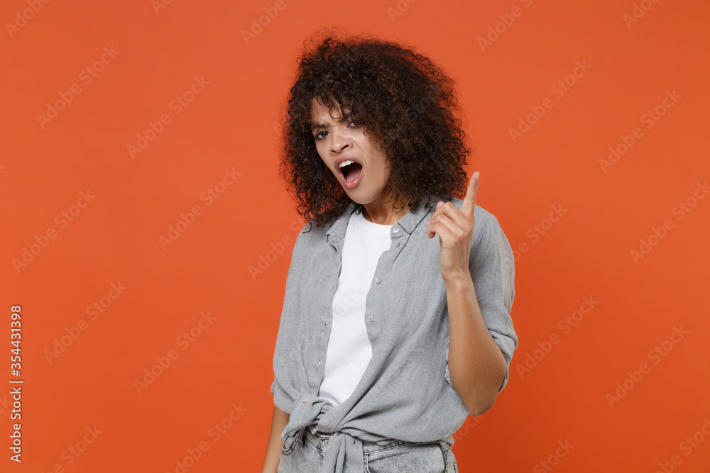 Displeased young african american woman girl in gray casual clothes isolated on orange wall background studio portrait. People lifestyle concept. Mock up copy space. Pointing index finger up swearing. Displeased young african american woman girl in gray casual clothes isolated on orange wall background studio portrait. People lifestyle concept. Mock up copy space. Pointing index finger up swearing.
