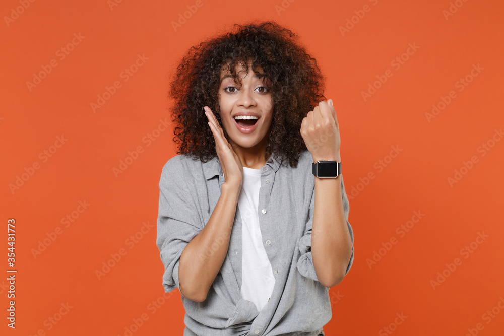 Excited young african american woman in gray casual clothes isolated on orange background studio portrait. People lifestyle concept. Mock up copy space. Wearing smart watch on hand put hand on cheek. Excited young african american woman in gray casual clothes isolated on orange background studio portrait. People lifestyle concept. Mock up copy space. Wearing smart watch on hand put hand on cheek.