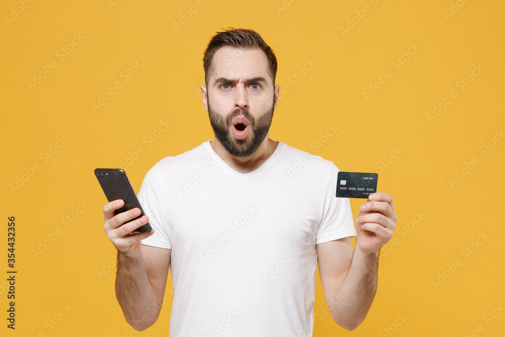 Shocked young bearded man guy in white casual t-shirt posing isolated on yellow wall background studio portrait. People lifestyle concept. Mock up copy space. Hold credit bank card using mobile phone. Shocked young bearded man guy in white casual t-shirt posing isolated on yellow wall background studio portrait. People lifestyle concept. Mock up copy space. Hold credit bank card using mobile phone.