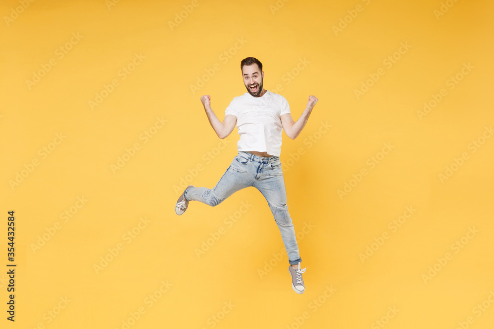 Excited young bearded man guy in white casual t-shirt posing isolated on yellow background studio portrait. People emotions lifestyle concept. Mock up copy space. Jumping clenching fists like winner.