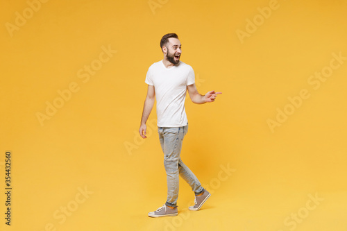 Excited young bearded man guy in white casual t-shirt posing isolated on yellow background studio portrait. People sincere emotions lifestyle concept. Mock up copy space. Pointing index finger aside.