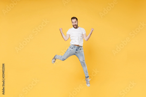 Excited young bearded man guy in white casual t-shirt posing isolated on yellow background studio portrait. People emotions lifestyle concept. Mock up copy space. Jumping clenching fists like winner.