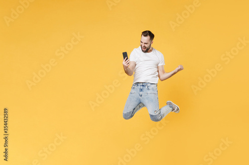 Happy young bearded man guy in white casual t-shirt posing isolated on yellow background. People lifestyle concept. Mock up copy space. Jumping doing selfie shot on mobile phone doing winner gesture.