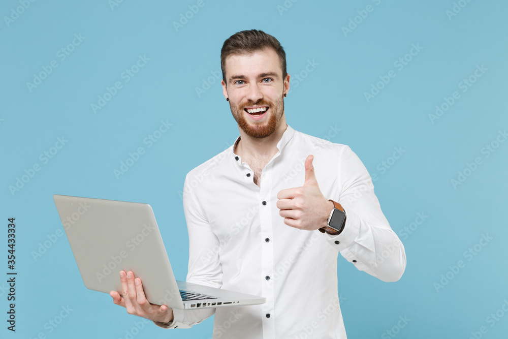 Cheerful young bearded man guy 20s in white classic shirt isolated on pastel blue wall background studio. People lifestyle concept. Mock up copy space. Working on laptop pc computer, showing thumb up. Cheerful young bearded man guy 20s in white classic shirt isolated on pastel blue wall background studio. People lifestyle concept. Mock up copy space. Working on laptop pc computer, showing thumb up.