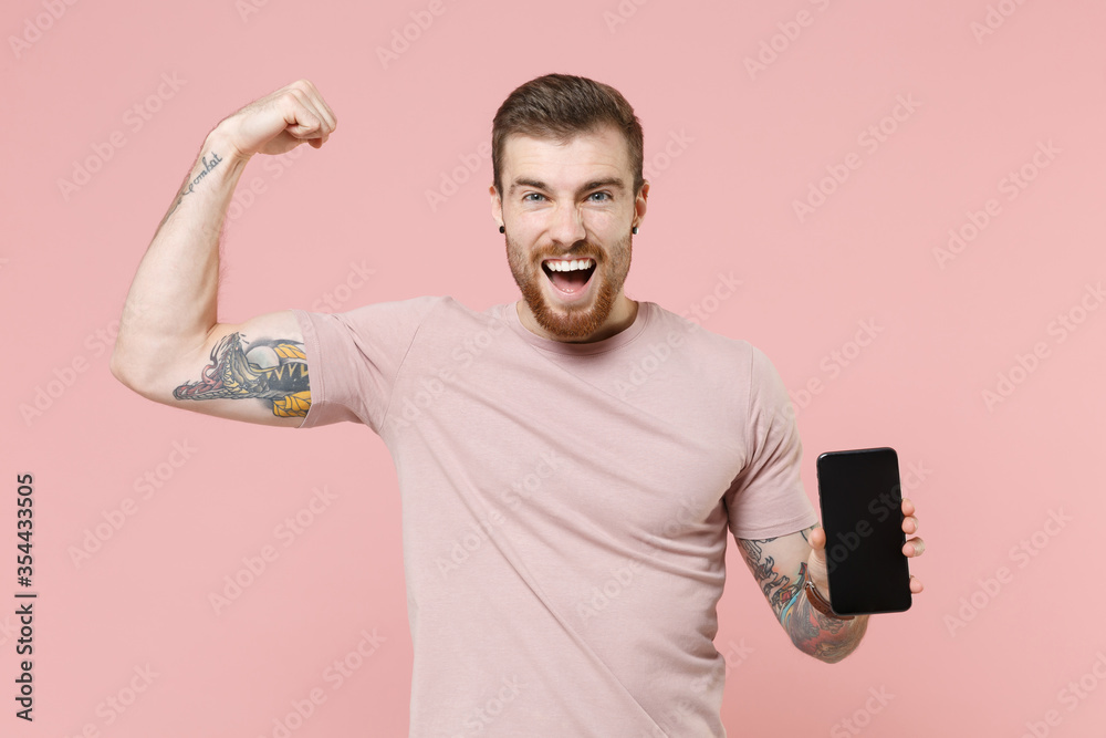 Joyful young bearded tattooed man guy in pastel t-shirt isolated on pink background. People lifestyle concept. Hold mobile phone with blank empty screen, showing biceps muscles doing winner gesture. Joyful young bearded tattooed man guy in pastel t-shirt isolated on pink background. People lifestyle concept. Hold mobile phone with blank empty screen, showing biceps muscles doing winner gesture.